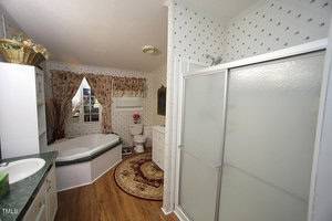 A bathroom with a corner bathtub, glass shower enclosure, toilet, white cabinets, a round rug, floral curtains on a window, and wallpaper with a small pattern. The floor is wood-style laminate.