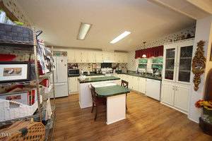 A spacious kitchen features white cabinets, green countertops, a central island with seating, wood flooring, open shelving on the left, a glass-front cabinet on the right, and a window with a red valance above the sink.
