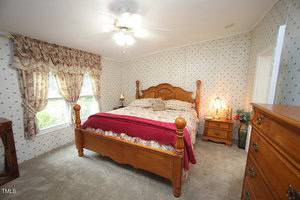 A bedroom with a wooden bed frame, a red and beige comforter, matching nightstand and dresser, a window with drapes, floral wallpaper, carpeted floor, and a ceiling fan with a light.