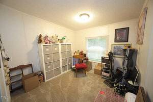 A home office or craft room with beige carpet, a filing cabinet with labeled bins, boxes, stuffed animals, an exercise machine, desk with computer equipment, and various decorations on the walls and shelves.