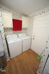 A laundry room with a white washing machine and dryer side by side, wooden floor, white cabinets, a red towel hanging between the cabinets, floral wallpaper, and a white door with a broom nearby.