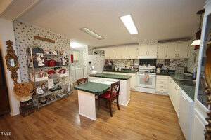 A spacious kitchen with white cabinets, green countertops, hardwood floors, a central island with two chairs, open shelving with dishes, and floral wallpaper. Fluorescent lights are on the ceiling.