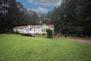 Located at 15 Cole Circle in Franklinton, this single-story white house with a red roof sits on a large grassy yard, featuring a covered carport to the right, a cozy front porch, and tall trees under a cloudy sky.