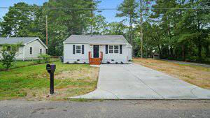 A small, single-story gray house with white trim and a black front door sits at the end of a wide concrete driveway. There is a black mailbox near the street, and the yard is bordered by trees and grass.
