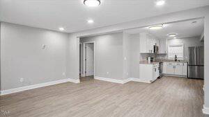 A modern open-plan living area with light wood flooring, white walls, recessed lighting, and a kitchen featuring white cabinets, granite countertops, stainless steel appliances, and a window above the sink.