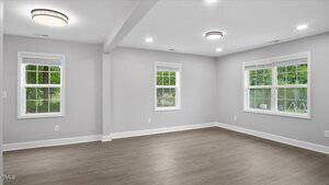 A spacious, empty room with light gray walls, large windows with white trim, recessed lighting, and wood-style flooring. Green trees and grass are visible through the windows.