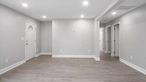 A room with light gray walls, recessed ceiling lights, wood-look flooring, a white front door, and a hallway leading to several white doors. The space is empty and clean.