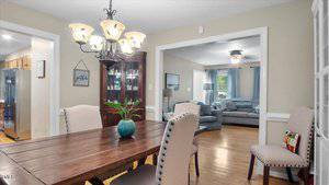 A dining room with a wooden table, four upholstered chairs, a hanging chandelier, and a teal vase with a plant; a living room with a gray sectional sofa is visible through a wide doorway.