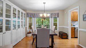 A dining room with a dark wooden table, six upholstered chairs, a glass-fronted white cabinet, a chandelier, a window with a valance, and an adjoining kitchen visible through an open doorway.