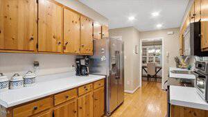 A kitchen with wood cabinets, white countertops, stainless steel appliances, a coffee maker, and decorative canisters. The floor is hardwood, and a dining area is visible through a doorway in the background.