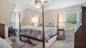 A bedroom with a four-poster bed covered in a patterned blue and white quilt, flanked by two bedside tables with lamps. There is a desk by the window, a ceiling fan, and neutral-colored walls and carpeting.