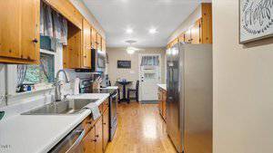A galley-style kitchen with wood cabinets, white countertops, stainless steel appliances, a large sink, and a small dining table near the back door. The floor is light wood and walls are beige.