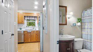 A view from a hallway shows a bathroom with a dark wood vanity and a white toilet on the right, and a kitchen with wood cabinets, stainless steel appliances, and a window on the left.