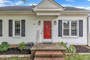 A white house with black shutters, a bright red front door, and a small porch with brick steps. There is a "WELCOME" sign by the door and plants on either side of the entry. House number 201 is visible next to the door.