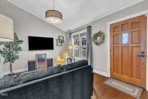 A living room with a black sofa, a wall-mounted TV, a yellow armchair, a potted plant, a floor lamp, a wooden front door, and a window with gray curtains. A wreath hangs on the wall by the door.