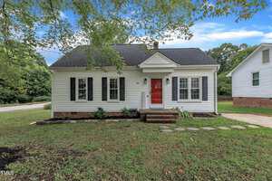 A small, single-story white house with black shutters, a red front door, and a gabled roof sits among green grass and trees at 201 Person Street in Oxford. A stone path leads to the entrance steps, and a driveway lines the right side.