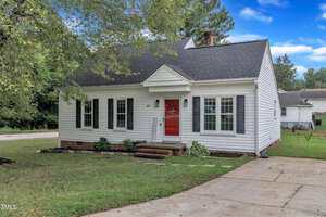A small white house with dark shutters and a red front door sits beside a driveway. There is a short brick walkway leading to the entrance, and a grassy yard with a few small plants and trees around the house.