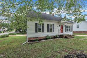 A small, single-story white house with black shutters and a red front door, set on a grassy lot with trees and a sidewalk. Brick steps lead to the entrance, and neighboring houses are visible nearby.
