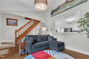 A living room with a dark gray sectional sofa, a colorful round rug, a wooden staircase, a potted plant, wall art, and a pass-through window showing a white kitchen in the background.