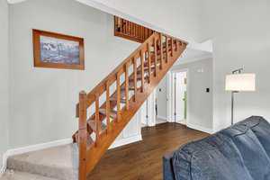 A wooden staircase with open risers leads to an upper level in a room with hardwood floors and light-colored walls. A framed painting hangs on the wall, and a blue couch is partially visible in the foreground.