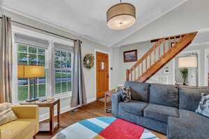 A living room with a gray couch, yellow armchair, round colorful rug, and wooden staircase. A wooden front door with a wreath and large window with gray curtains are visible. A lamp sits on a small table by the window.