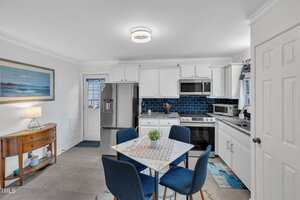 A modern kitchen with white cabinets, stainless steel appliances, blue tile backsplash, and a small dining table with four blue chairs. A side table with a lamp and a framed ocean picture is on the left.
