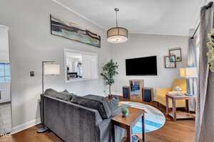 Modern living room with a gray sectional sofa, round rug, large wall-mounted TV, yellow armchair, wooden side tables, floor lamp, potted plant, abstract wall art, and bright natural light from windows.