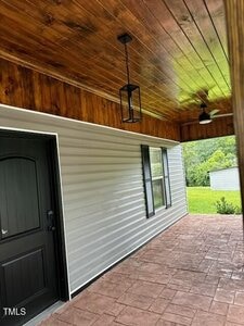 Covered porch area with a wood-paneled ceiling, square light fixture, ceiling fan, black door, two windows, and textured floor. Green grass and a small building are visible in the background.