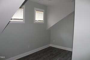 A small, empty room with light gray walls, dark wood flooring, two small windows with blinds, and sloped ceilings creating angular shapes on the left and right sides.