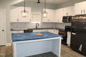 A modern kitchen with white cabinets, stainless steel appliances, marble backsplash, and two pendant lights over a center island. The island is covered in blue protective plastic. A paper sits on the island.