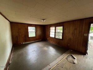 Empty room with wood-paneled walls, two windows, a ceiling light fixture, and a bare floor. The baseboard heater is below one window, and some floor covering is missing near the entrance.
