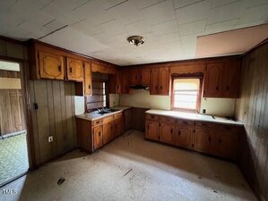 A dimly lit, outdated kitchen with wood-paneled walls, old wooden cabinets, a sink under a small window, and a stained ceiling. The floor appears worn and the space is empty, with no appliances or furniture.