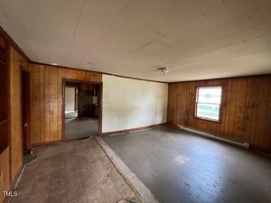 An empty room with wood-paneled walls, a single window, and a bare floor. The ceiling is white with a central light fixture. An adjacent room is visible through an open doorway.