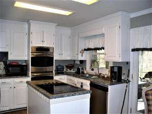 A kitchen with white cabinets, stainless steel double ovens, a cooktop on a center island, a dishwasher, a coffee maker, and a window above the sink. The room is well-lit with fluorescent ceiling lights.