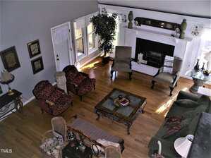 A living room with hardwood floors, a fireplace, several chairs, a sectional sofa, a central coffee table, a side table with a lamp, framed pictures on the wall, and a potted plant near a window.