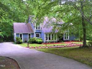 A two-story blue house with white trim is surrounded by trees and greenery. A curved concrete driveway leads to the front of the house, with landscaped beds and a well-maintained lawn in the yard.