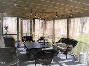 A screened porch with wicker chairs, a cushioned sofa, a central square fire pit table, overhead string lights, wind chimes, and views of trees outside. The space appears bright with natural light.