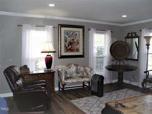 A living room with gray walls, wood floor, a floral loveseat, brown leather recliner, wooden desk with a red lamp, wall art, a small table with a decorative plate, mirror, floor lamp, and portable heater.