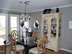 A dining room with a glass table, four wooden chairs, a white china cabinet displaying glassware, a chandelier, a round mirror on the wall, and two windows letting in natural light.