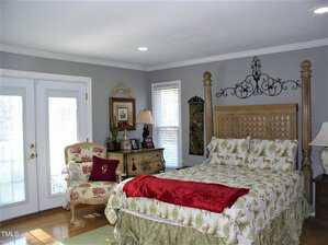 A bedroom with a large wooden bed, floral bedding, and a red throw blanket. There is an upholstered chair and a dresser with a lamp. French doors and two windows let in natural light. The walls are painted light gray.