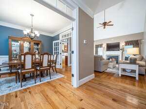 Spacious home interior showing a dining area with a wooden table and chairs, a living room with sofas, a coffee table, lamp, ceiling fan, fireplace, hardwood floors, and large windows with curtains.