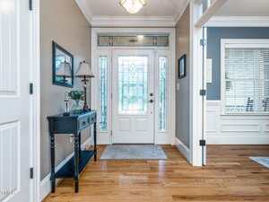 A bright entryway with a white front door featuring decorative glass panels, a narrow black console table with a lamp and decor, light gray walls, wood flooring, and crown molding.
