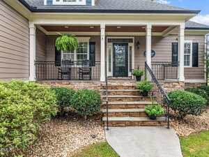 A front porch with two black rocking chairs, potted plants, a hanging fern, and stone steps leading to a glass front door. The house has beige siding, white trim, and landscaped bushes along the walkway.