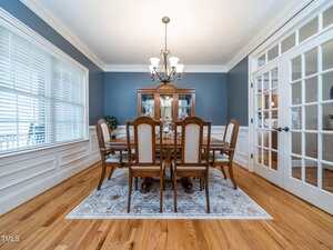 A formal dining room with a wooden table and eight chairs on a patterned rug, a chandelier overhead, a glass-fronted cabinet against a blue wall, and double glass doors to the right. White trim and hardwood floors are visible.