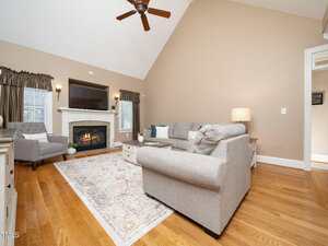 A living room with wood floors, a gray sectional sofa, a fireplace with a TV above it, two armchairs, an area rug, a ceiling fan, and windows with dark curtains on either side of the fireplace.