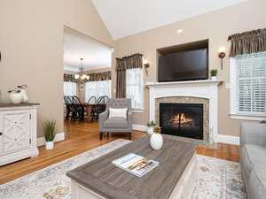 A living room with a lit fireplace, a TV above the mantel, an armchair with a "Home" pillow, a coffee table with magazines and a small pineapple decor, and dining area visible in the background.