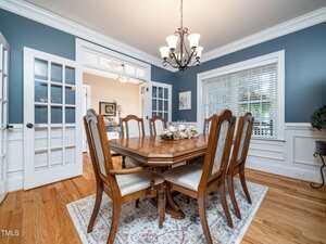A dining room with a wooden table and eight chairs, a decorative centerpiece, a rug underneath, hardwood floors, blue walls with white trim, a window with blinds, and a chandelier overhead.