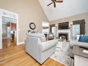 A spacious living room with light gray sofas, a white coffee table, a fireplace with a mantel, hardwood floors, and a ceiling fan. The area leads to a dining space and kitchen, with large windows letting in natural light.