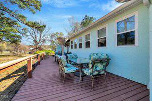 A spacious wooden deck with a glass-top table and six cushioned chairs is attached to a light blue house. Trees, a railing, and clear blue skies are visible in the background.