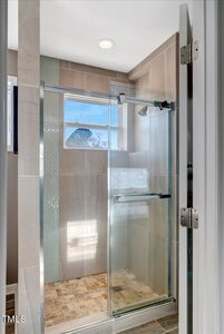 A modern walk-in shower with a glass door, tiled walls in neutral tones, a vertical accent of small green tiles, and a window letting in natural light above the shower area.